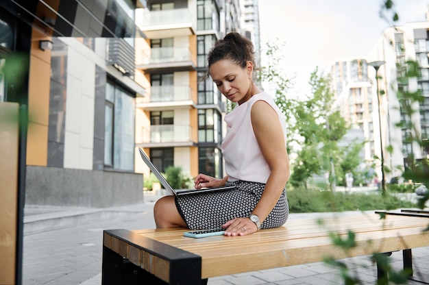 Young African American business woman working on laptop, looking at screen of a smartphone on urban background . Female entrepreneur using laptop and chatting. Woman freelancer portrait.