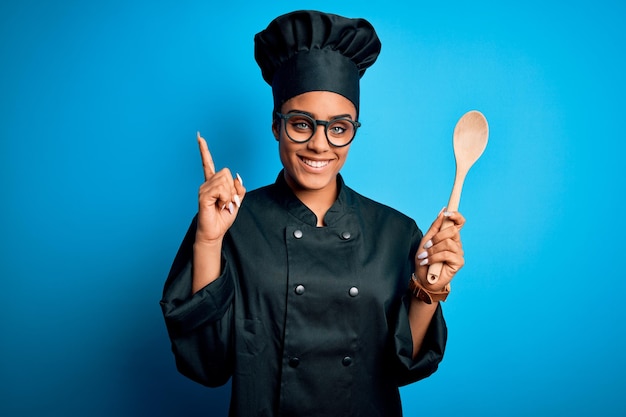 Young african american chef girl wearing cooker uniform and hat holding wooden spoon tool surprised with an idea or question pointing finger with happy face number one