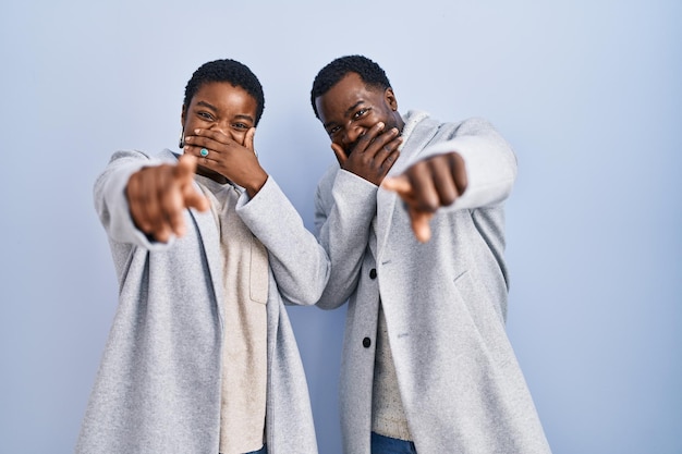 Photo young african american couple standing over blue background together laughing at you pointing finger to the camera with hand over mouth shame expression