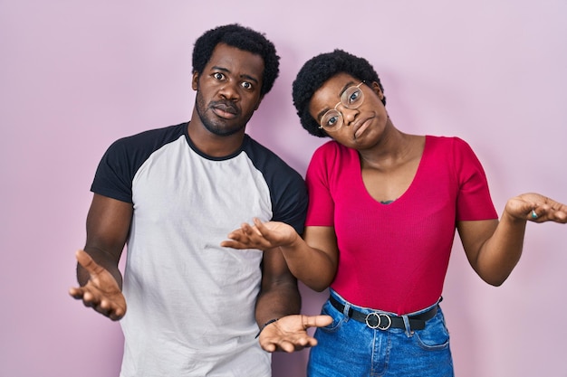 Young african american couple standing over pink background clueless and confused with open arms, no idea concept.