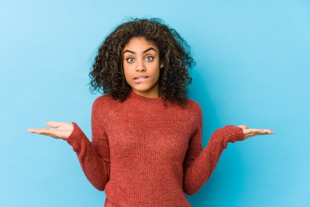 Young african american curly hair woman confused and doubtful shrugging shoulders to hold a copy space