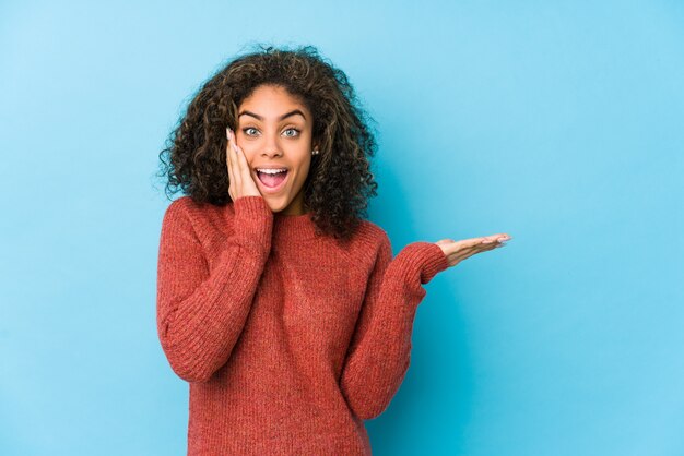 Young african american curly hair woman holds blank space on a palm, keep hand over cheek.