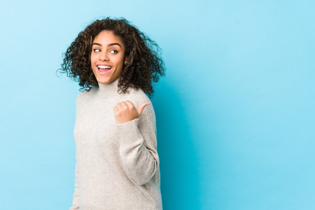 Young african american curly hair woman points with thumb finger away, laughing and carefree.