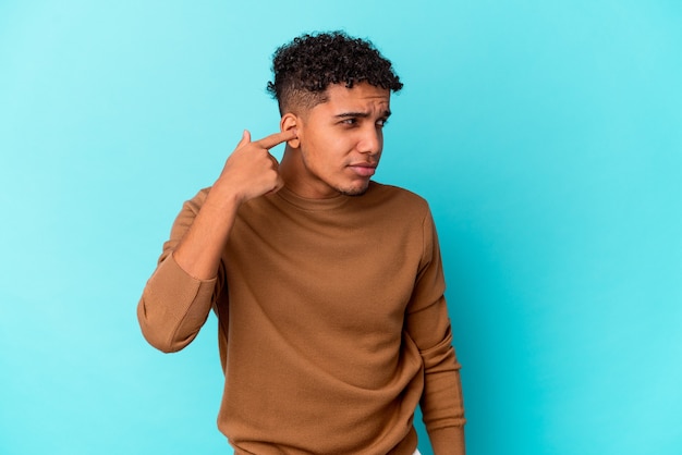 Young african american curly man isolated on blue covering ears with fingers, stressed and desperate by a loudly ambient.