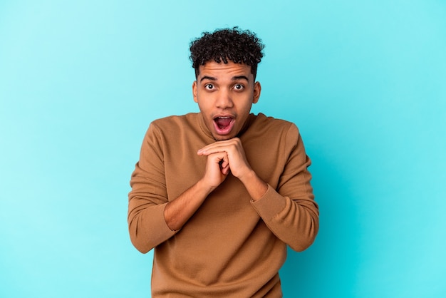 Young african american curly man isolated on blue praying for luck, amazed and opening mouth looking to front.