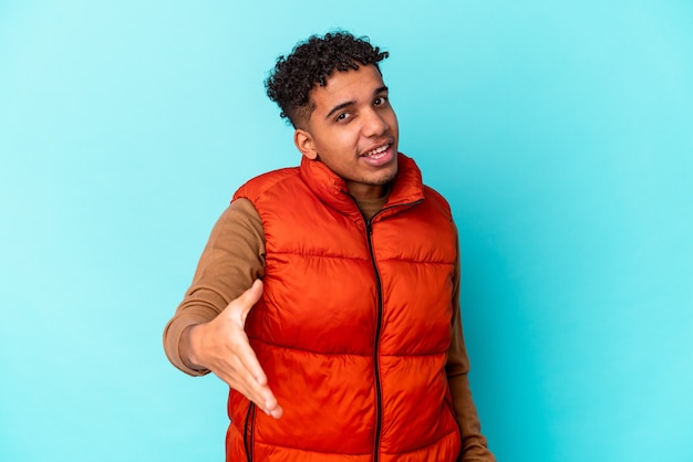 Young african american curly man isolated on blue stretching hand at camera in greeting gesture.