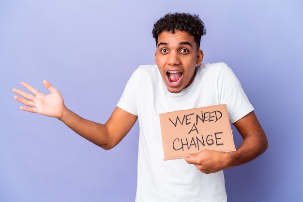 Young african american curly man isolated holding a we need a change cardboard receiving a pleasant surprise, excited and raising hands.