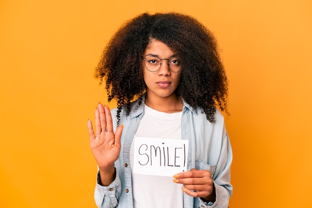 Young african american curly woman holding a smile message placard standing with outstretched hand showing stop sign, preventing you.