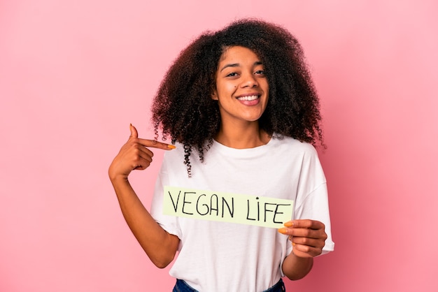 Young african american curly woman holding a vegan life placard person pointing by hand to a shirt copy space, proud and confident