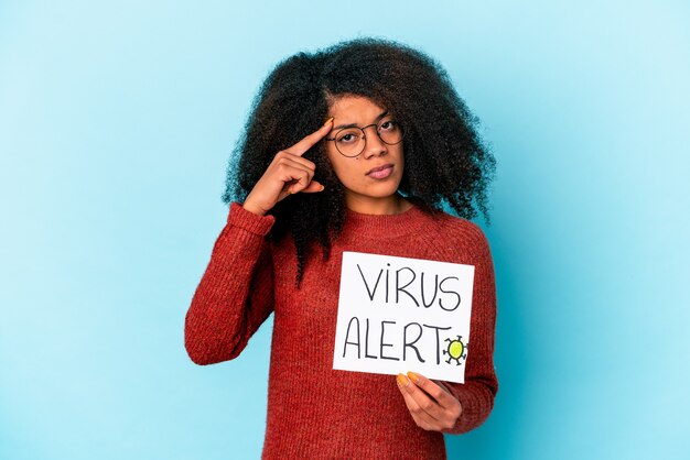 Young african american curly woman holding a virus alert placard pointing temple with finger, thinking, focused on a task.