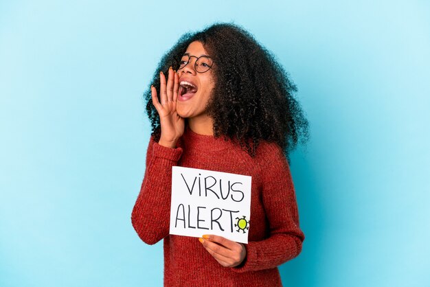 Young african american curly woman holding a virus alert placard shouting and holding palm near opened mouth.