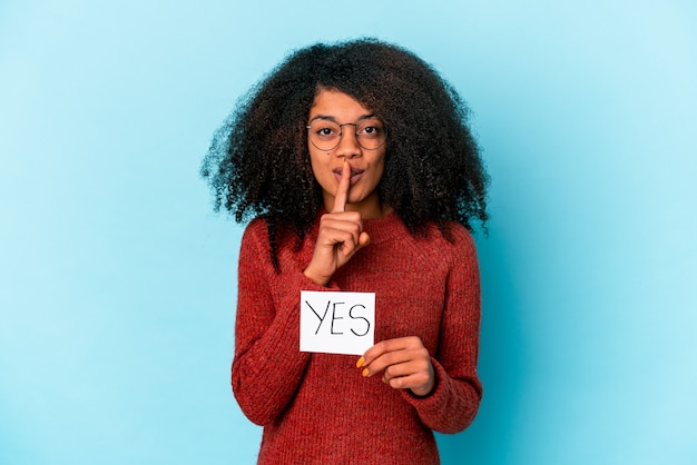 Young african american curly woman holding a yes placard keeping a secret or asking for silence.