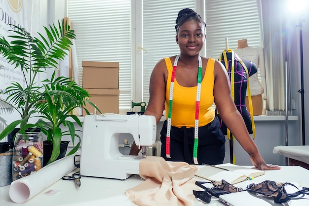 Young african american dressmaker woman sews clothes on sewing machine at tailor office it tropical summer style