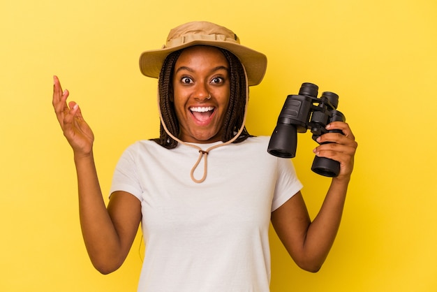 Young african american explorer woman holding a binoculars isolated on yellow background  receiving a pleasant surprise, excited and raising hands.