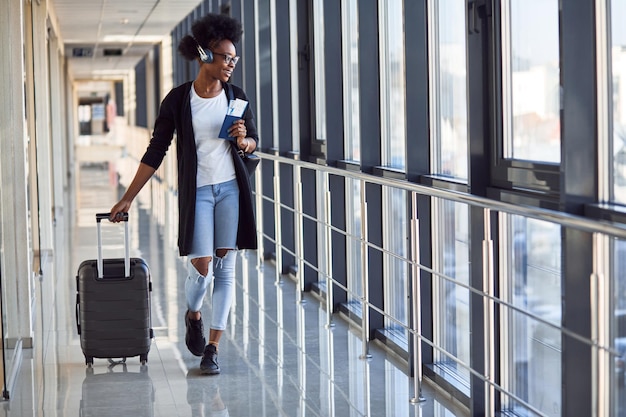 Young african american female passanger in casual clothes and headphones is in airport with baggage.