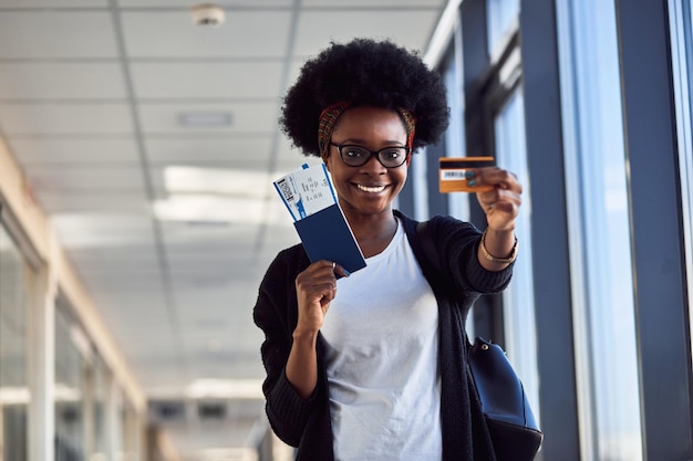 Young african american female passanger in casual clothes is in airport holding tickets and credit card.