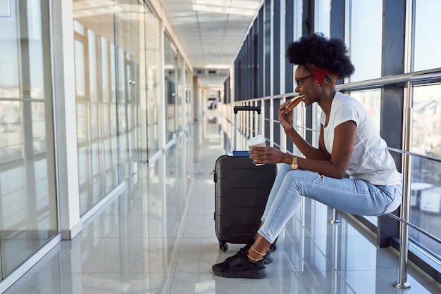 Il giovane passeggero femminile afroamericano in abiti casual è in aeroporto con il bagaglio che mangia del cibo.