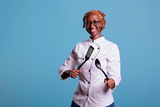 Young african american female restaurant crew member posing with kitchen utensils. Curly haired chef portrait smiling, holding a spoon, spatula. Kitchen helper laughing.