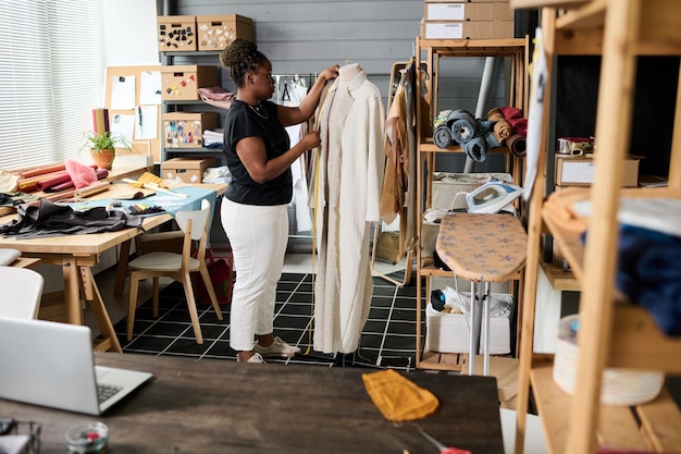 Young African American female tailor standing in front of mannequin with coat on