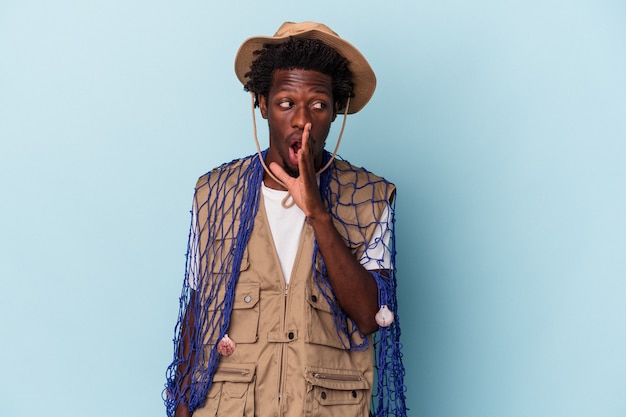 Young african american fisherman holding a net isolated on blue background is saying a secret hot braking news and looking aside