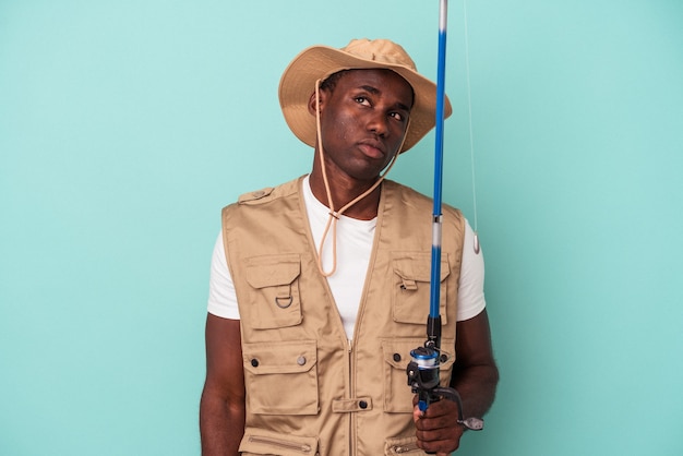 Young African American fisherman holding rod isolated on blue background dreaming of achieving goals and purposes