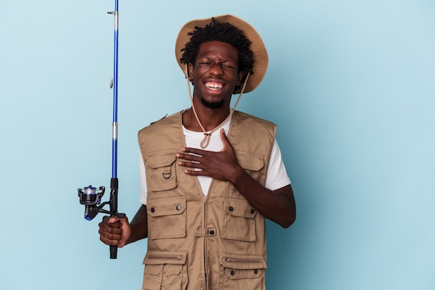 Young african american fisherman holding a rod isolated on blue background laughs out loudly keeping hand on chest.