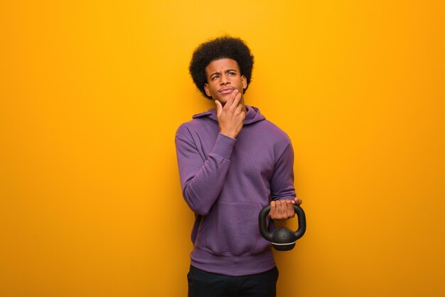 Young african american fitness man holding a dumbbell doubting and confused