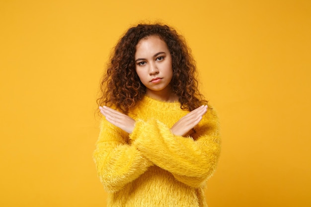 Young african american girl in fur sweater posing isolated on yellow orange wall background studio portrait. People lifestyle concept. Mock up copy space. Showing stop gesture with crossed hands.