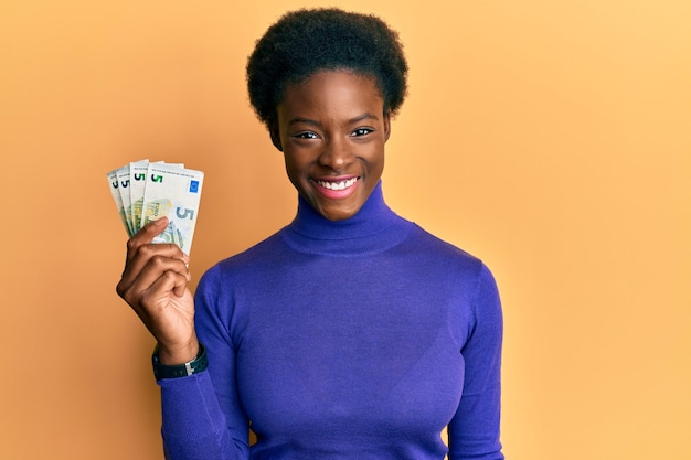 Young african american girl holding bunch of 5 euro banknotes looking positive and happy standing and smiling with a confident smile showing teeth