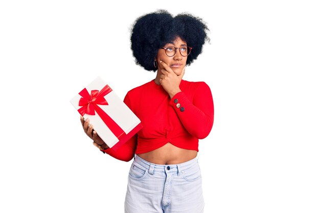 Young african american girl holding gift thinking worried about a question concerned and nervous with hand on chin