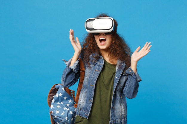 Young african american girl teen student in denim clothes, backpack headset vr isolated on blue wall