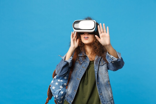 Young african american girl teen student in denim clothes, backpack headset vr isolated on blue wall