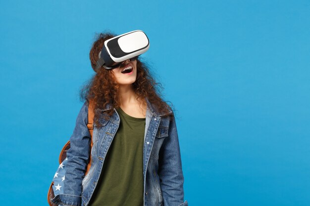 Young african american girl teen student in denim clothes, backpack headset vr isolated on blue wall