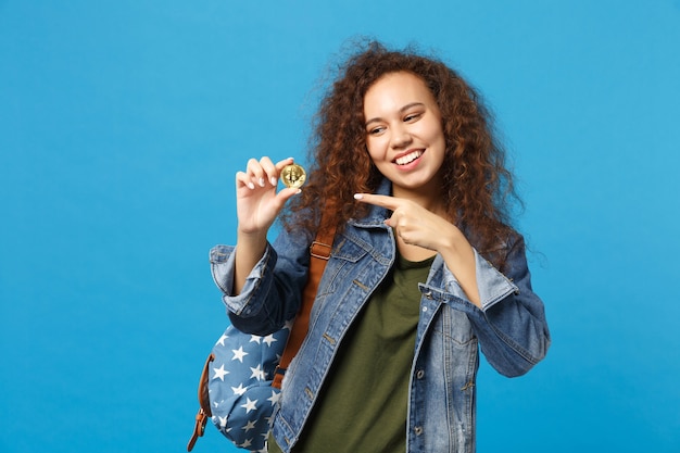 Young african american girl teen student in denim clothes, backpack hold bitcoin isolated on blue wall