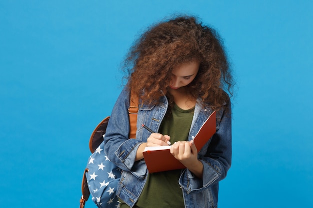 Giovane studentessa teenager della ragazza afroamericana in vestiti del denim, libri della tenuta dello zaino isolati sulla parete blu