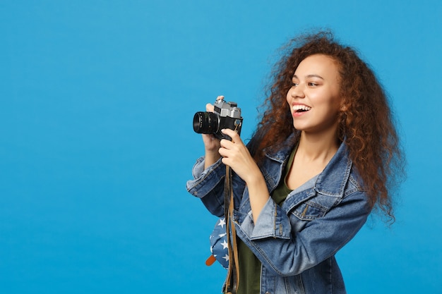 Young african american girl teen student in denim clothes backpack hold camera isolated on blue wall