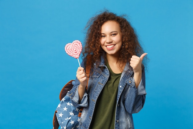 Young african american girl teen student in denim clothes, backpack hold candy isolated on blue wall