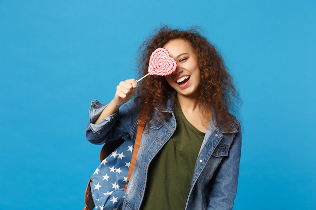 Young african american girl teen student in denim clothes, backpack hold candy isolated on blue wall