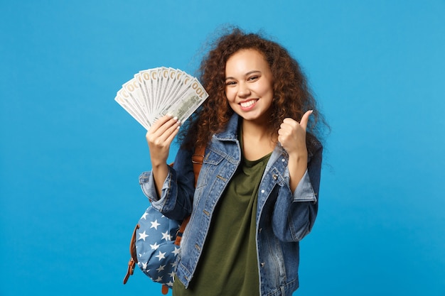 Young african american girl teen student in denim clothes, backpack hold cash money isolated on blue wall