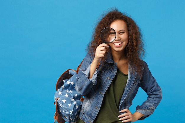 Молодая афро-американская девушка-подросток-студент в джинсовой одежде, рюкзак держит часы, изолированные на синей стене