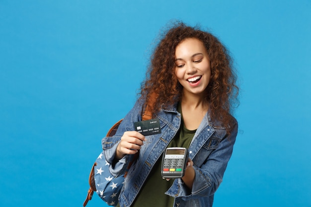 Young african american girl teen student in denim clothes, backpack hold credit card isolated on blue wall