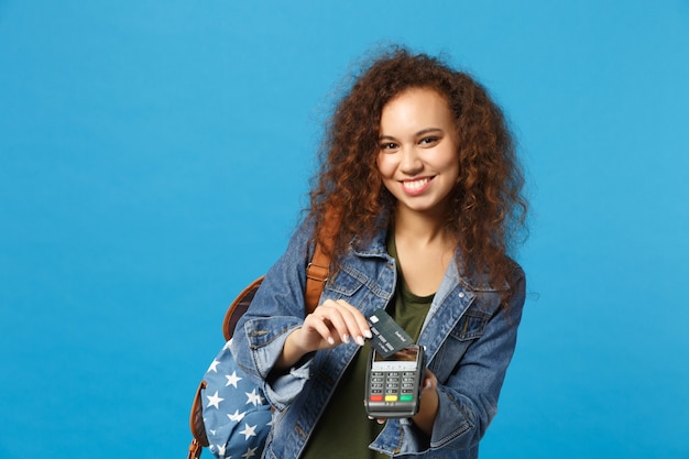 Young african american girl teen student in denim clothes, backpack hold credit card isolated on blue wall