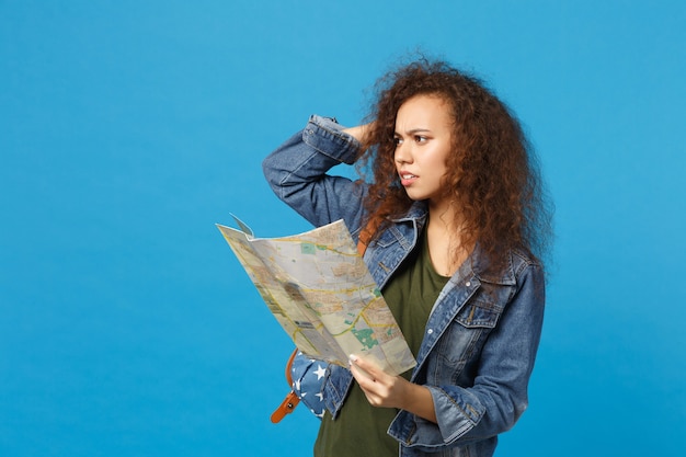 Young african american girl teen student in denim clothes, backpack hold map isolated on blue wall