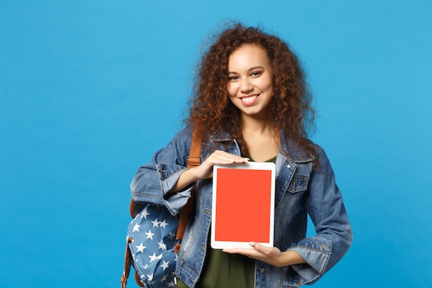 Young african american girl teen student in denim clothes, backpack hold pad pc isolated on blue wall wall