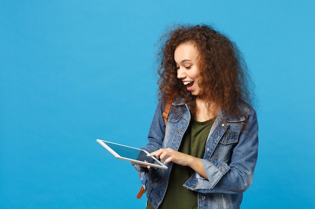 Young african american girl teen student in denim clothes, backpack hold pad pc isolated on blue wall wall