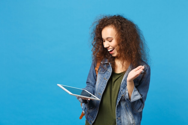 Young african american girl teen student in denim clothes, backpack hold pad pc isolated on blue wall wall