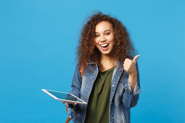 Young african american girl teen student in denim clothes, backpack hold pad pc isolated on blue wall wall