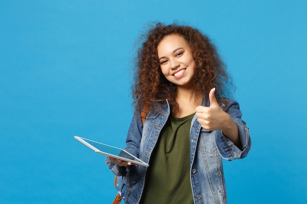 Young african american girl teen student in denim clothes, backpack hold pad pc isolated on blue wall wall