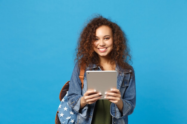 Young african american girl teen student in denim clothes, backpack hold pad pc isolated on blue wall wall