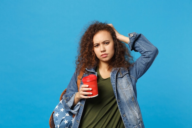 Giovane studentessa teenager della ragazza afroamericana in vestiti del denim, tazza di carta della tenuta dello zaino isolata sulla parete blu